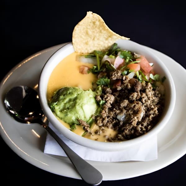 Queso dip with ground beef, guacamole, pico de gallo, and a tortilla chip in a white bowl.