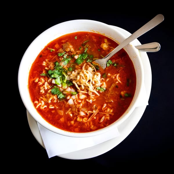 A white bowl of red ramen soup with cilantro, onions, and meat, with a fork inside.