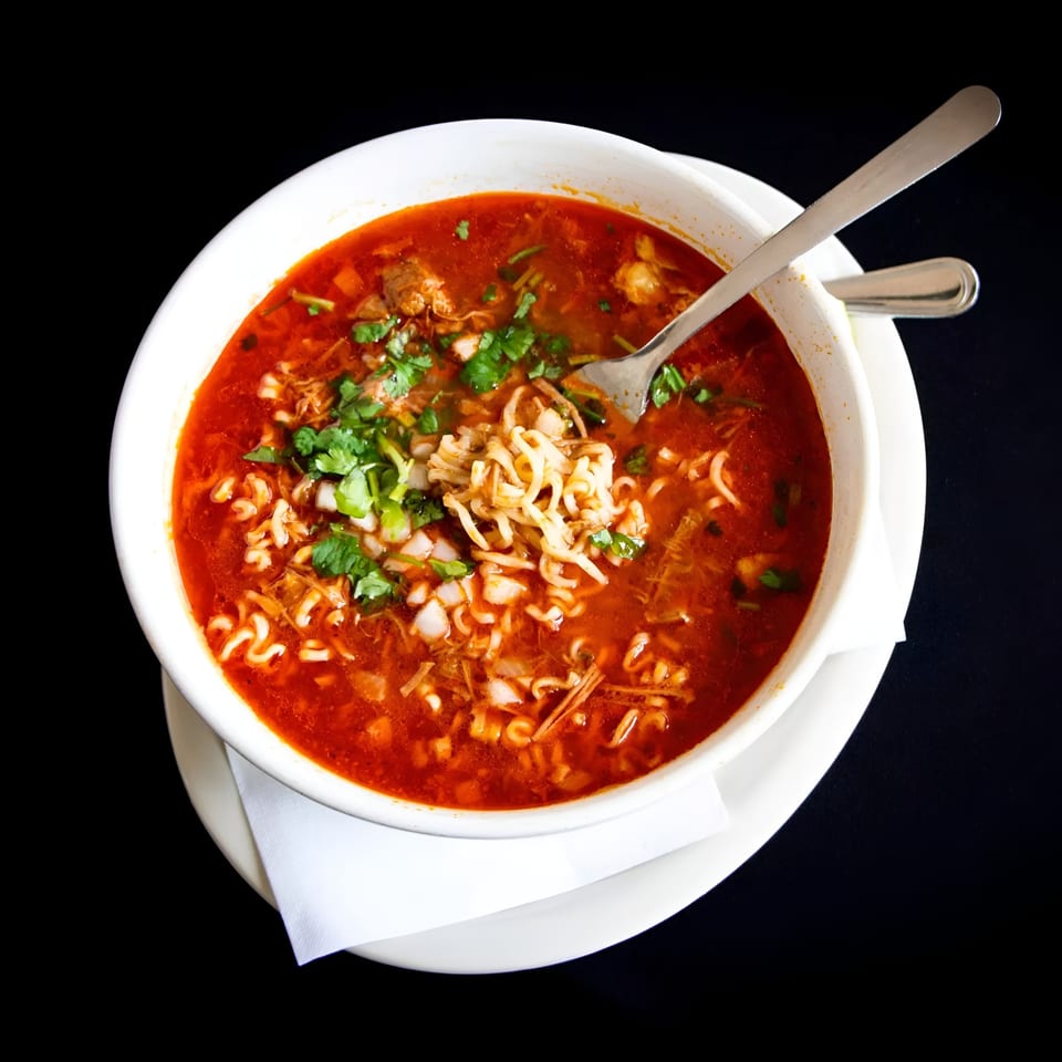 A white bowl of red ramen soup with cilantro, onions, and meat, with a fork inside.