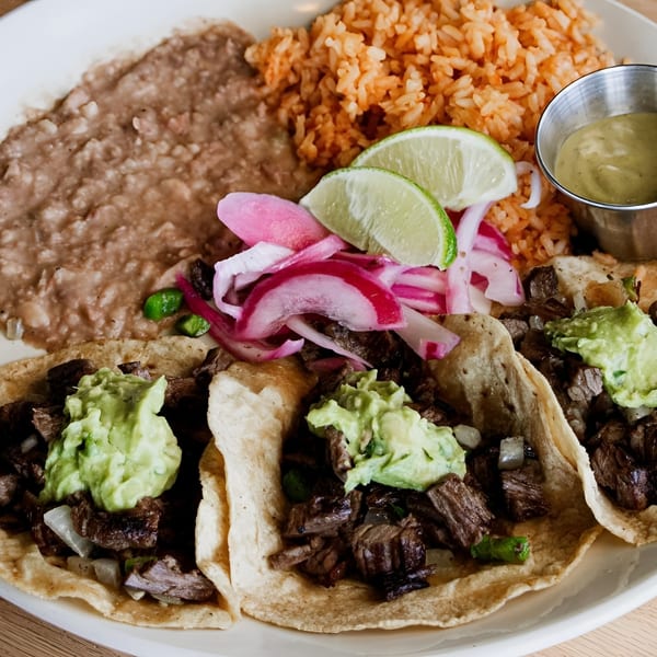 Three street tacos with guacamole, pickled onions, lime, rice, and refried beans on a white plate.