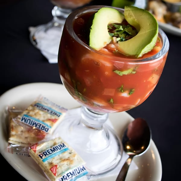 Shrimp cocktail in a glass with avocado slices and cilantro, served with crackers and a spoon.