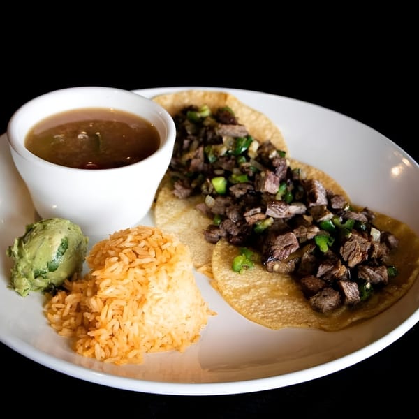 Two beef tacos with rice, guacamole, and a cup of broth on a white plate.