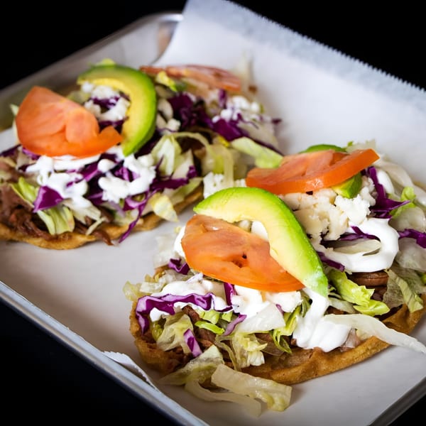 Two sopes topped with shredded meat, lettuce, purple cabbage, sour cream, crumbled cheese, tomato, and avocado.