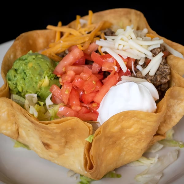 A taco salad in a crispy tortilla bowl with ground beef, cheese, tomatoes, guacamole, and sour cream.